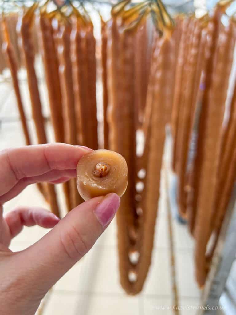 A hand holds a sliced piece of churchkhela, a traditional Georgian candy with a nut center, with whole churchkhela strands hanging in the background.