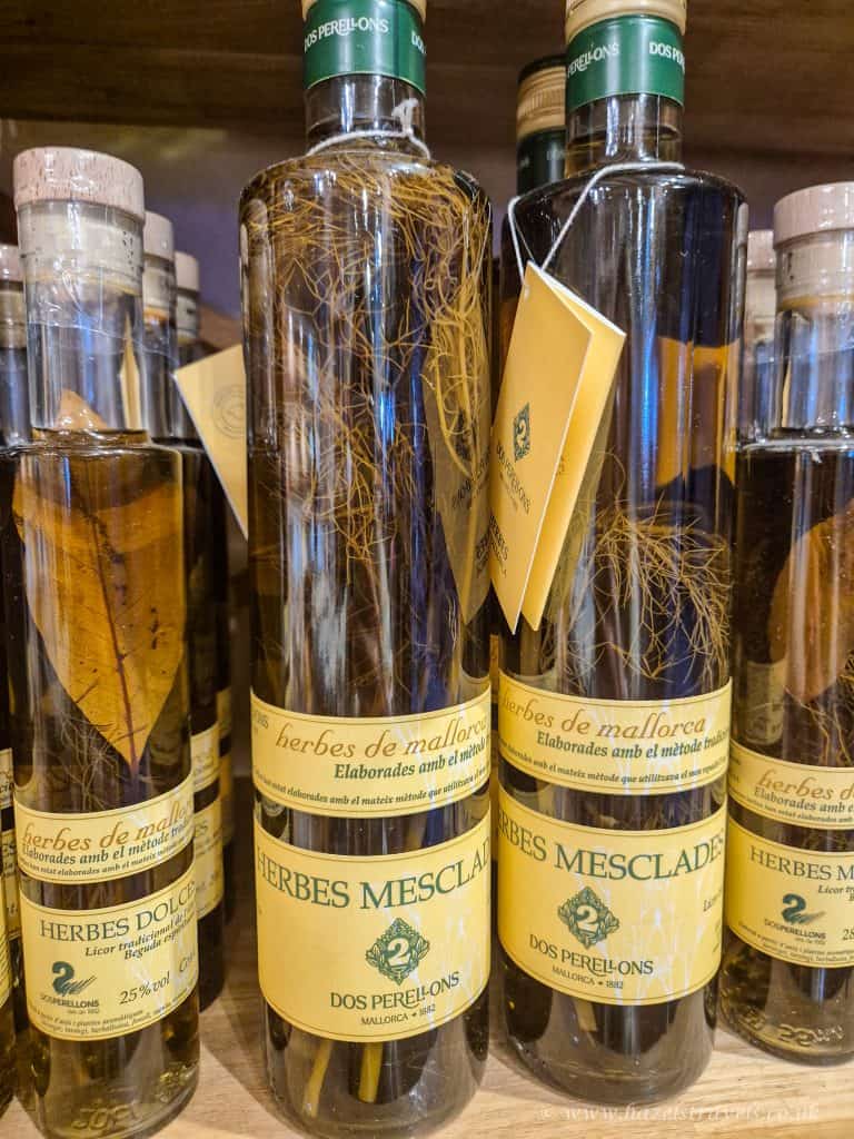 Bottles of Herbes de Mallorca liquor labeled “Herbes Mesclades” and “Herbes Dolces,” with visible herbs and leaves inside, displayed on a wooden shelf.