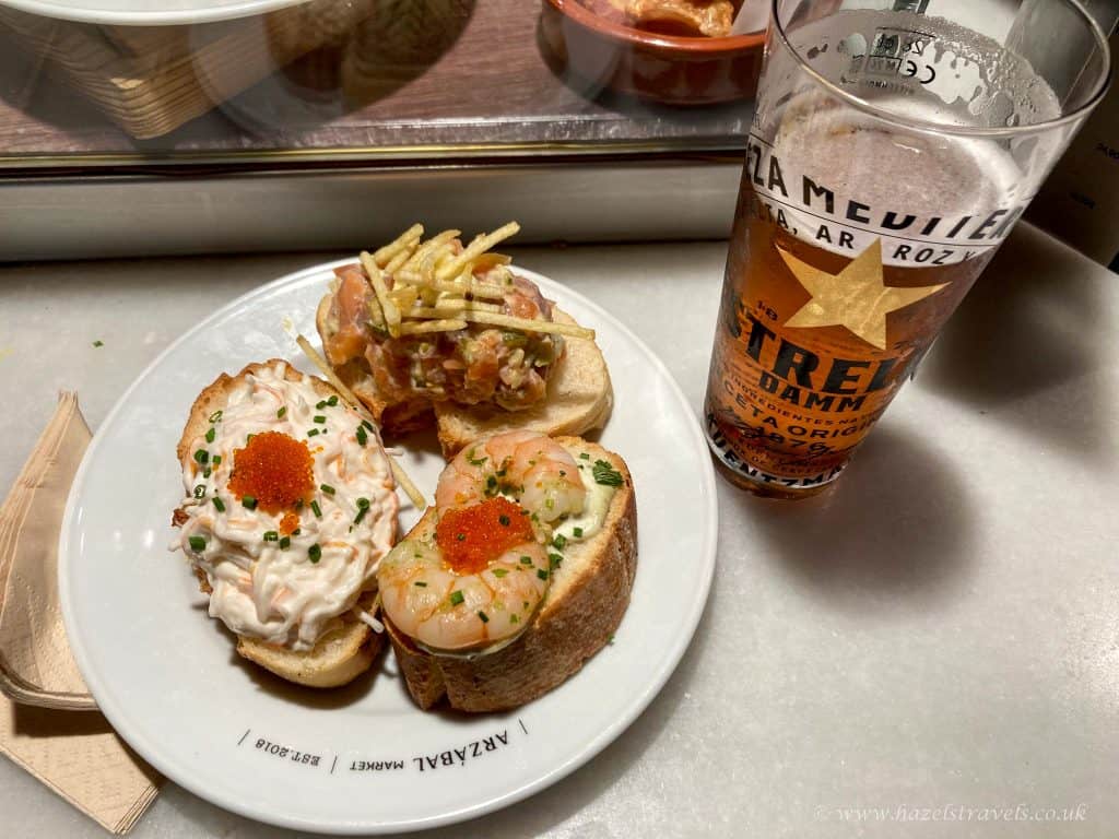 Delicious tapas and a cold beer served on a plate, showcasing Spanish cuisine. Pastries topped with seafood, cream, and garnishes, with a refreshing beer in the background.