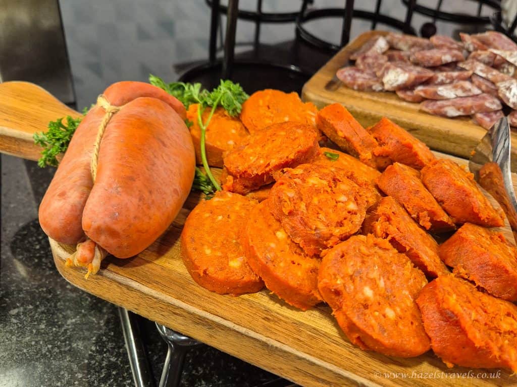 A wooden board displays slices of bright orange sobrasada sausage next to a whole sausage and garnished with parsley; sliced cured meats are in the background.