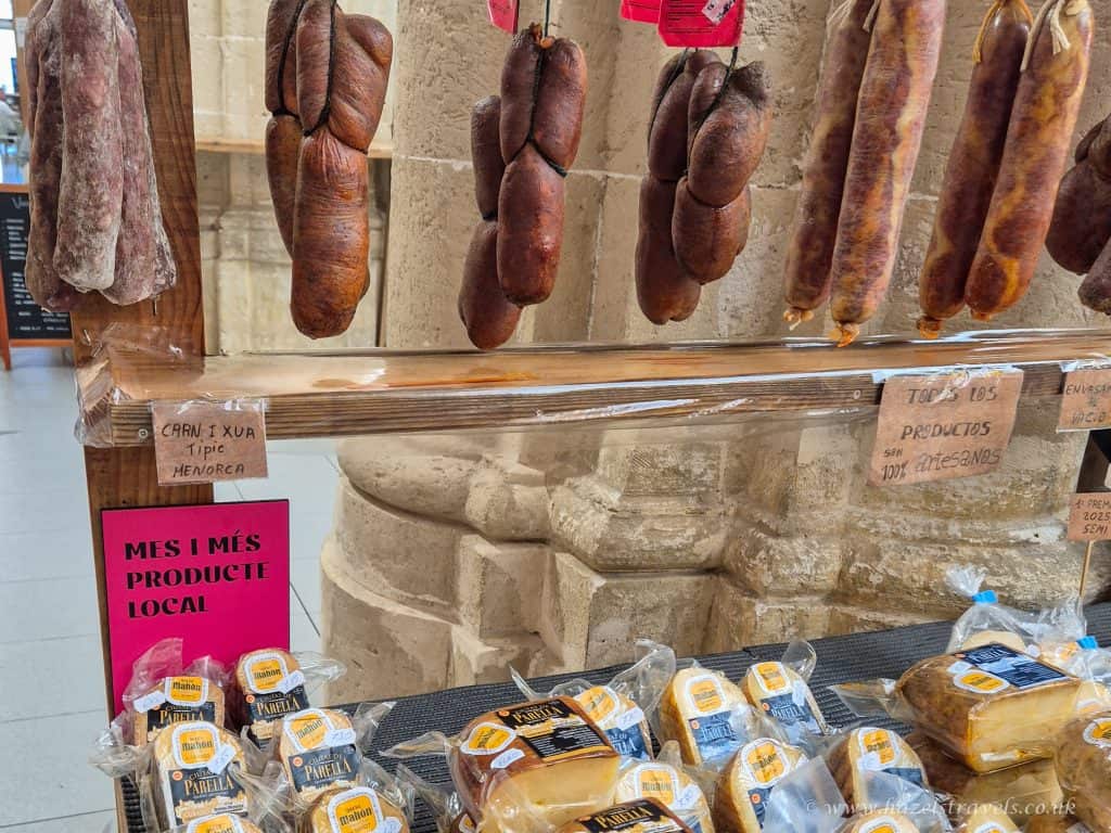 Various types of cured sausages hang above a wooden shelf displaying packaged local cheese and bread in a Menorca market setting.