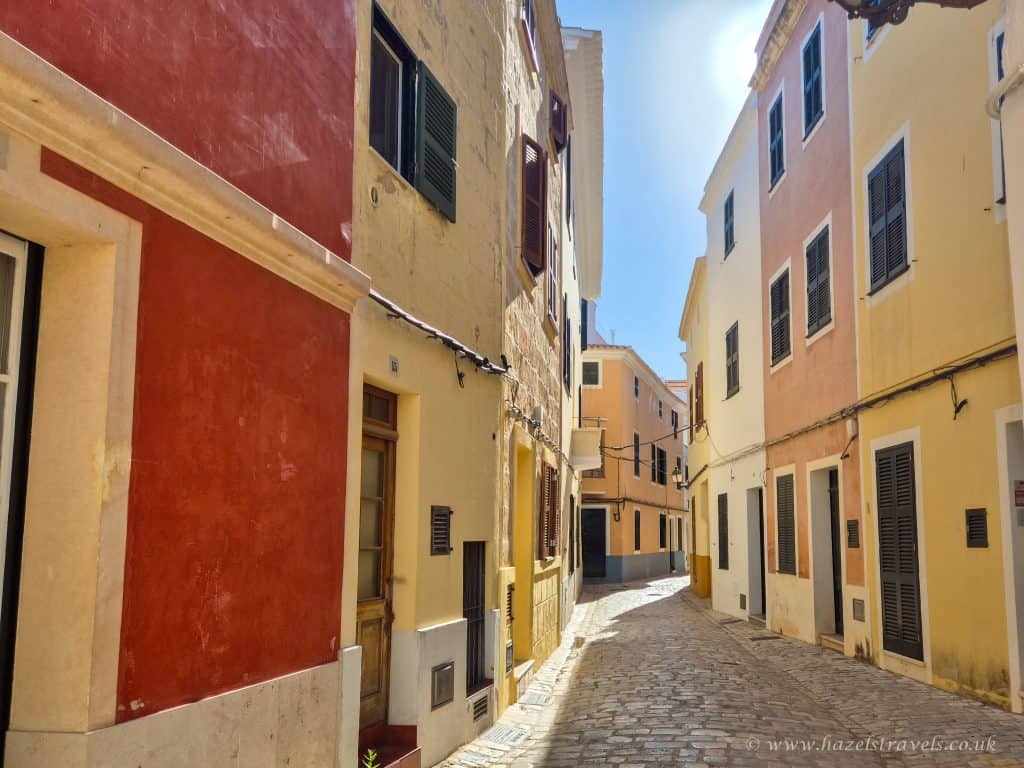 Colourful Mediterranean alleyway with narrow cobblestone street, vibrant pastel buildings, and sunny sky.