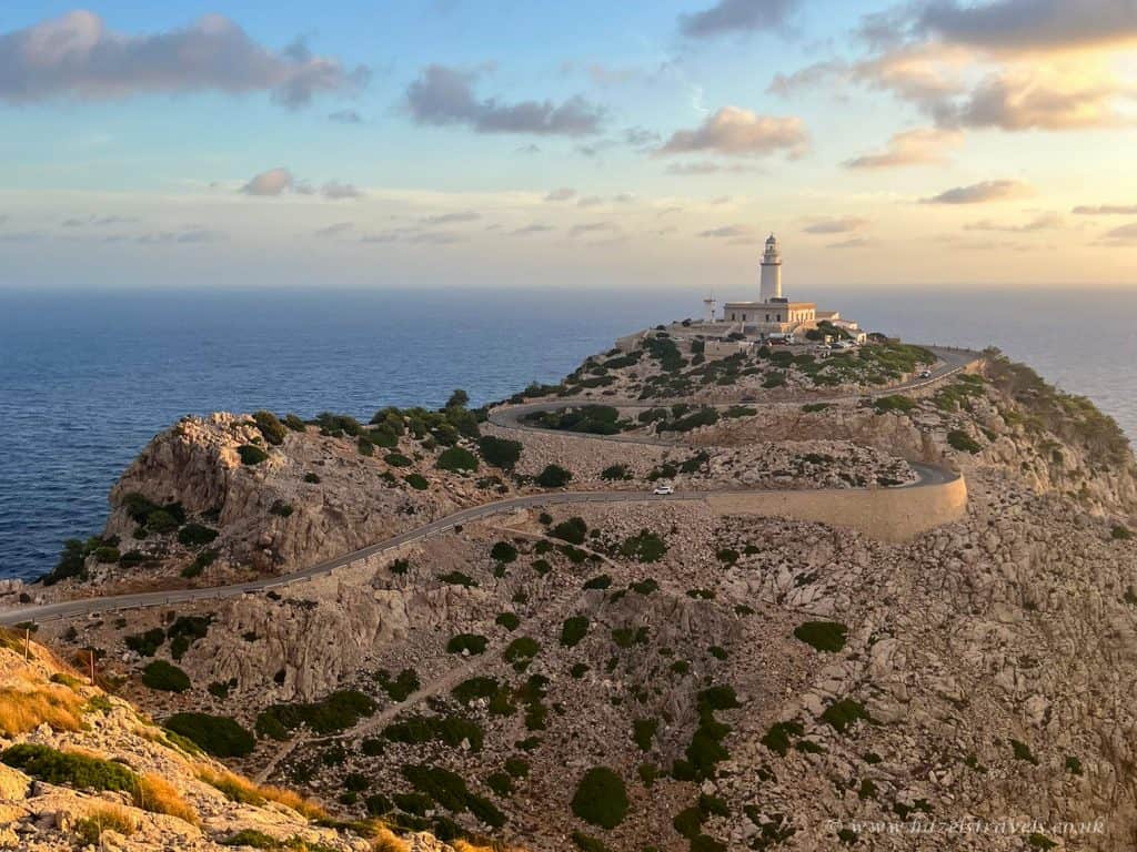 A lighthouse stands on a rocky cliff above a winding coastal road, overlooking the sea under a partly cloudy sky at sunset.