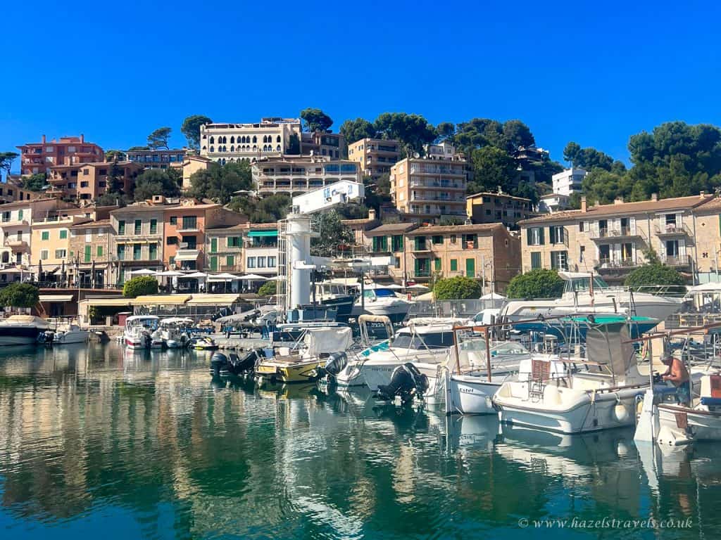Boats are docked in a marina with colorful buildings and green trees on a hillside in the background under a clear blue sky.