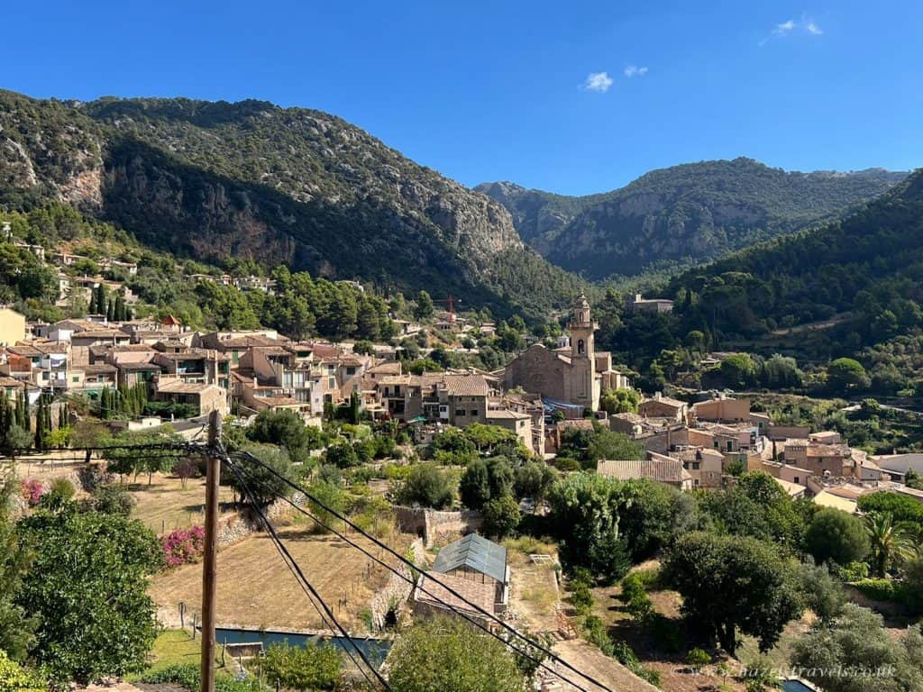 A small village with stone buildings and a central church is nestled among green hills and mountains under a clear blue sky.